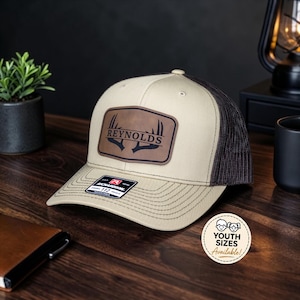 May include: A beige and dark brown trucker hat featuring a rectangular brown leather patch on the front. The patch displays the text REYNOLDS above a graphic of deer antlers. A black sticker is on the brim, and a circular graphic states YOUTH SIZES Available.