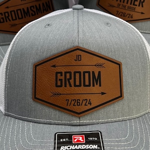 May include: A gray and white mesh baseball cap with a brown leather patch that says "JD GROOM 7/26/24". The patch has two arrows pointing in opposite directions. The cap has a black "R" logo on the front.