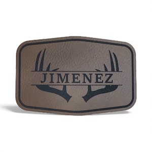 May include: A rectangular brown leather patch with rounded corners and a black border. The patch features a black graphic of deer antlers with a horizontal line running through the center, containing the black text JIMENEZ in capital letters.
