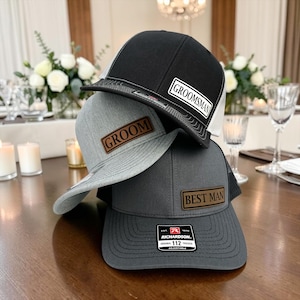 May include: Three adjustable trucker hats stacked together. The top hat is black with a white patch reading GROOMSMAN. The middle hat is heather gray with a brown leather patch reading GROOM. The bottom hat is dark gray with a brown patch reading BEST MAN.