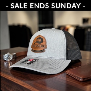 May include: A gray trucker hat with a black mesh back and a leather patch. The patch features the text "YOUR LOGO HERE" and a logo design. The hat is on a wooden surface.