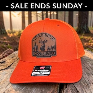 May include: A bright orange trucker hat featuring a brown leather patch with a deer head graphic, pine trees, and the text LITTLE BUCK SHOOTER CLUB LIFETIME MEMBER. A black sticker on the brim reads RICHARDSON 112 TRUCKER EST 1970.