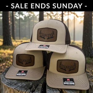 May include: Three tan and brown trucker hats with leather patches featuring the names "Lassiter" and "McGregor" and a deer antler design. The hats have a mesh back and a curved brim. The text "SALE ENDS SUNDAY" is at the top.