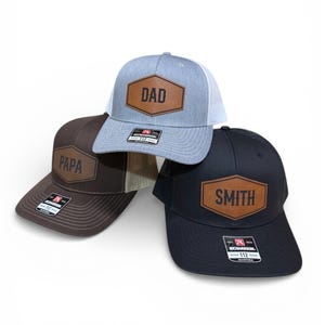 May include: Three adjustable trucker hats featuring a hexagonal brown leather patch on the front. The light gray hat displays the text DAD, the dark brown hat displays the text PAPA, and the black hat displays the text SMITH. Each hat has a Richardson 112 label.