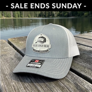 May include: A gray and white trucker hat with a patch that says "YOUR LOGO HERE". The hat has a curved brim and a mesh back. The text "- SALE ENDS SUNDAY -" is at the top of the image. The hat is adjustable.