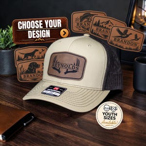 May include: A beige and dark brown trucker hat featuring a rectangular leather patch with the name REYNOLDS and an antler graphic. The hat has a structured front, a mesh back, and a Richardson 112 sticker on the brim. Additional leather patch designs are displayed in the background.