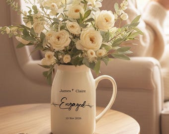 May include: A white ceramic vase with a handle, filled with white roses and greenery, sits on a wooden table. The vase is inscribed with "James + Claire Engaged 10 Nov 2026". A ring rests on the table. A person is seated in a chair in the background.
