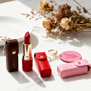 Puede incluir: Una colección de pintalabios y estuches en varios colores. El pintalabios es rojo con un tubo dorado. Los estuches son marrones, rojos y rosas, con llaveros dorados. Un llavero rosa tiene el nombre "Celine".