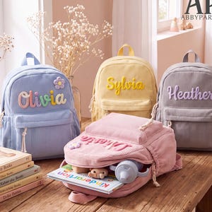 May include: Four corduroy backpacks in light blue, yellow, gray, and pink. Each bag features a name embroidered in raised letters on the front pocket. The blue and pink bags include a small flower applique. One pink bag is open, revealing a book.