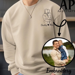 May include: A beige sweatshirt with a black line drawing of a father and daughter with the text "Daddy Mia" underneath. The sweatshirt is shown with an embroidered image of a father and daughter in a circle with the word "Embroidery" underneath.