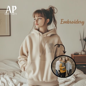 Peut inclure: Un sweat à capuche beige avec un dessin au trait noir de trois personnes sur le devant. Le sweat à capuche a une capuche et une poche kangourou. Le texte "Embroidery" est écrit en brun sur le côté droit de l'image. Une photo de trois personnes est représentée dans un cercle sur le côté droit de l'image.
