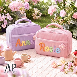 May include: Two rectangular corduroy lunch bags with top handles and zippered closures. One bag is lavender with the name Anne in colorful yarn letters, and the other is pink with the name Rosie in colorful yarn letters, each featuring a small flower.