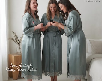 Puede incluir: Tres mujeres con batas verde polvoriento con ribete de encaje, sonriendo y mirando un teléfono. Las batas tienen cierre de lazo y llegan a la rodilla. El texto "New Arrival: Dusty Green Robes" es visible.