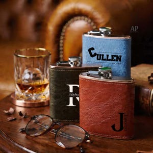 Pode incluir: Três cantis de couro personalizados em azul, verde e castanho, cada um com uma inicial e um nome únicos. Um copo de whisky e um par de óculos estão em primeiro plano. Os cantis têm uma tampa de aço inoxidável.