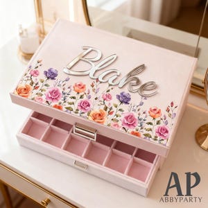 May include: A light pink jewelry box with the name "Blake" in silver lettering on the lid. The lid is decorated with a floral design featuring pink, purple, and orange roses. The open box reveals a pink interior with multiple compartments.