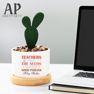 Op de afbeelding: Een witte keramische plantenpot met een houten basis, met een groene cactusplant. De pot heeft de tekst "Teachers Plant The Seeds That Will Grow Forever Miss Bella" in roze en oranje letters.