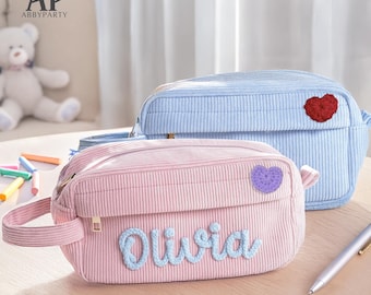 May include: A light pink corduroy pencil case featuring the name Olivia in blue script lettering and a small purple crocheted heart patch, positioned in front of a light blue corduroy pencil case with a red crocheted heart patch.