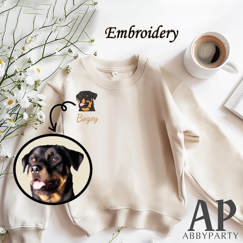 May include: A beige sweatshirt with an embroidered image of a Rottweiler dog with the name "Bogey" underneath. The sweatshirt is on a white surface with a cup of coffee and a white frame.