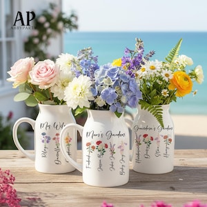 Op de afbeelding: Drie witte keramische vazen met handvatten, elk gevuld met kleurrijke bloemen. De vazen zijn gelabeld met "Mrs. Wife", "Mum's Garden" en "Grandma's Garden" met namen eronder. De bloemen omvatten rozen, madeliefjes en hortensia's.