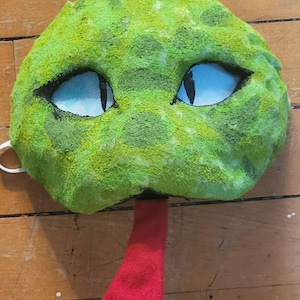 May include: A handmade green snake mask with blue eyes and a red tongue. The mask is textured with a spotted pattern and has a white elastic band. Perfect for costume parties or imaginative play.