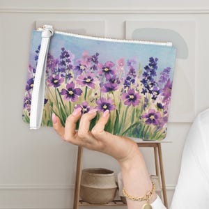 May include: A floral clutch with a watercolor design featuring purple flowers and green grass against a light blue sky. The clutch has a white wrist strap and a gold zipper. The overall theme is nature and art.