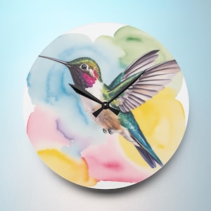 May include: Round wall clock featuring a watercolor hummingbird with outstretched wings. The bird has green, red, and blue plumage, set against a backdrop of colorful watercolor washes in yellow, pink, and blue. The clock hands are black.