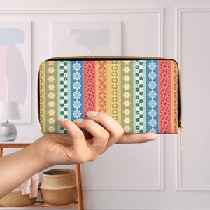 May include: A colorful floral pattern wallet with a black zipper. The wallet features vertical stripes in various colors, including yellow, green, blue, and red, adorned with flower and dot designs. The wallet is held in a person's hand.