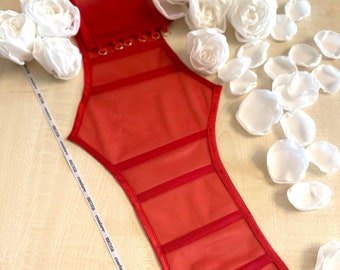 Könnte beinhalten: Rotes Netzkorsett mit goldenen Ringen und einer roten Satinband-Schleife. Das Korsett besteht aus einem durchsichtigen Netzstoff mit rotem Satinbesatz. Das Korsett liegt auf einer weißen Oberfläche mit weißen Blütenblättern.