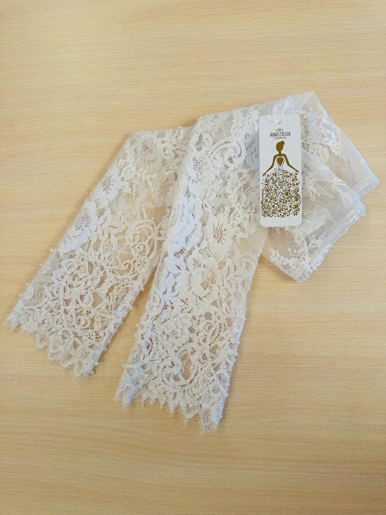 Peut inclure: Gants de mari&eacute;e en dentelle blanche avec des embellissements de perles. Les gants sont pli&eacute;s en deux et portent une petite &eacute;tiquette blanche avec le texte "Anastasia Bridal".