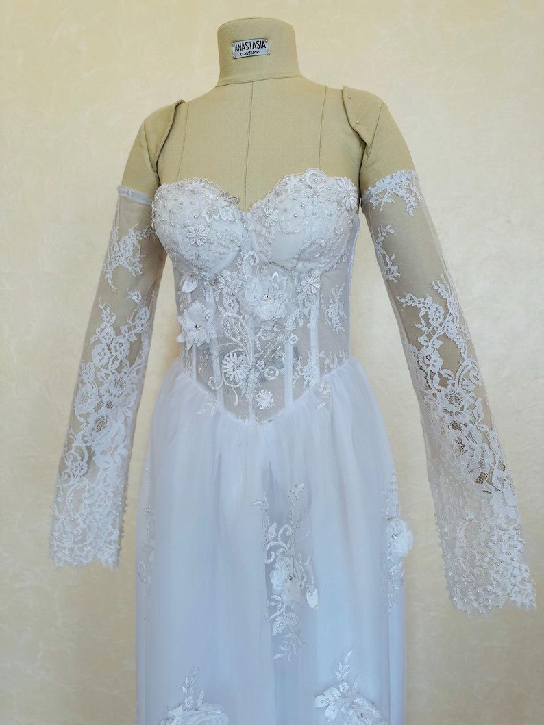 Peut inclure: Une robe de mari&eacute;e en dentelle blanche avec un corsage ajust&eacute; et une jupe ample. La robe a des manches longues avec des d&eacute;tails en dentelle et un d&eacute;collet&eacute; en c&oelig;ur.