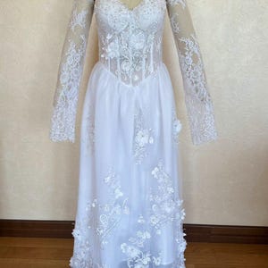 Peut inclure: Une robe de mari&eacute;e blanche avec de longues manches en dentelle et un corsage en dentelle. La jupe est en tulle et est brod&eacute;e de fleurs. La robe est sur un mannequin.