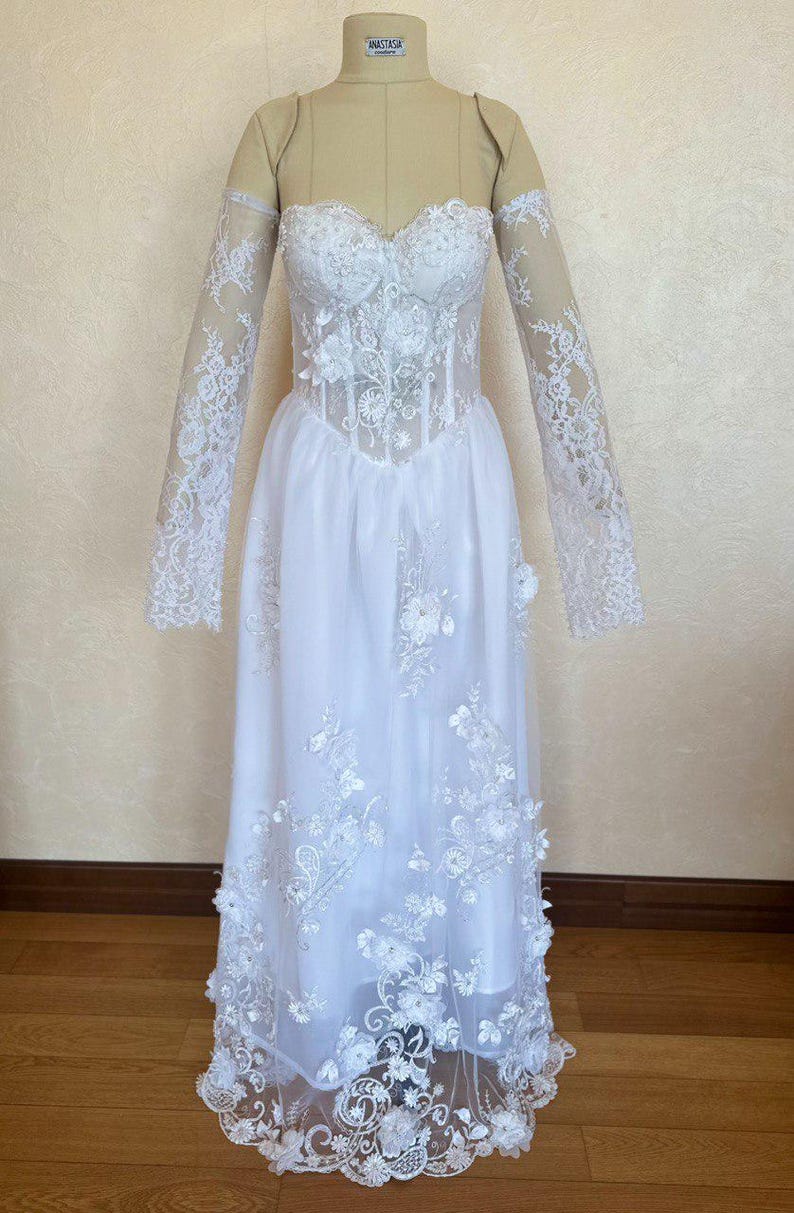 Peut inclure: Une robe de mari&eacute;e blanche avec de longues manches en dentelle et un corsage en dentelle. La jupe est en tulle et est brod&eacute;e de fleurs. La robe est sur un mannequin.
