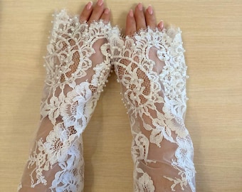 May include: A pair of white fingerless lace gloves featuring intricate floral patterns and small white pearl embellishments. The gloves extend from the knuckles to the mid-forearm, showcasing a delicate sheer mesh base with scalloped edges.