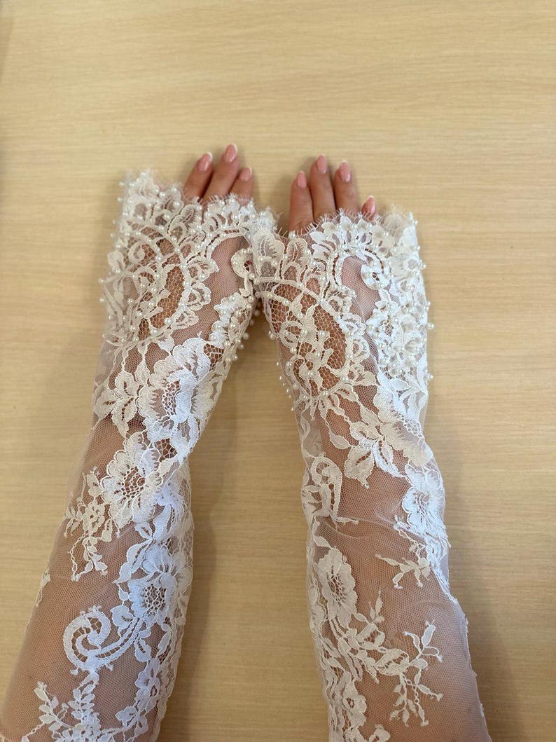 Peut inclure: Une paire de gants en dentelle blanche avec des embellissements de perles. Les gants sont longs et atteignent le coude. La dentelle est d&eacute;licate et complexe, et les perles sont espac&eacute;es uniform&eacute;ment.