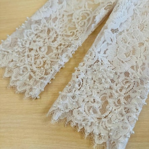 Peut inclure: Gros plan sur le bord inf&eacute;rieur de deux manches en dentelle blanche avec une rang&eacute;e de perles blanches cousues le long du bord.
