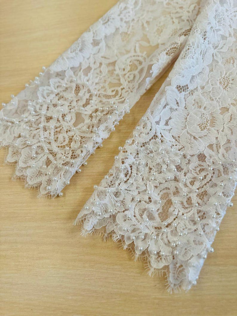 Peut inclure: Gros plan sur le bord inf&eacute;rieur de deux manches en dentelle blanche avec une rang&eacute;e de perles blanches cousues le long du bord.