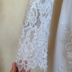 Peut inclure: Tissu de dentelle blanche avec des embellissements de perles. La dentelle a un motif floral et les perles sont cousues autour des bords.