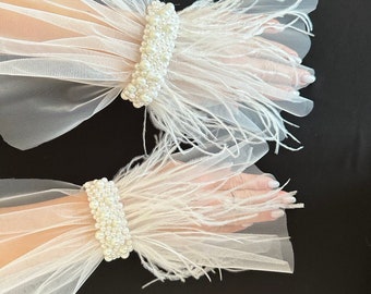 May include: White tulle wrist cuffs with pearl accents and feather trim. The cuffs are designed with multiple layers of sheer fabric, creating a delicate, airy look. The cuffs are adorned with small, round pearls.