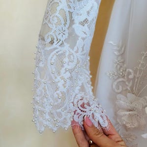 Peut inclure: Robe de mari&eacute;e en dentelle blanche avec des embellissements de perles sur les manches. La robe a un corsage ajust&eacute; et une jupe ample.