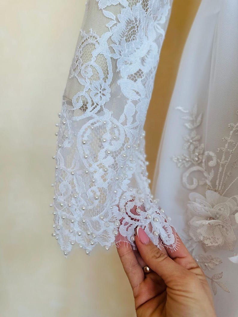 Peut inclure: Robe de mari&eacute;e en dentelle blanche avec des embellissements de perles sur les manches. La robe a un corsage ajust&eacute; et une jupe ample.