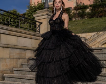 May include: A black formal gown with a tiered tulle skirt and halter neckline. The dress features intricate detailing on the bodice and is worn with strappy heels. The background includes a building and a staircase.