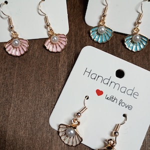 May include: Three pairs of dangle earrings on white display cards labeled Handmade with love. Each earring features a gold-tone metal shell charm with enamel in pink, light blue, or white, accented with a small white faux pearl at the center.