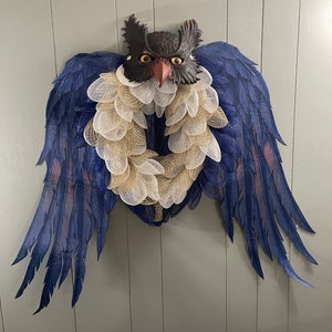 May include: A decorative owl wreath with a dark brown owl head and large blue wings. The wreath features a gold and white mesh design, creating a unique and eye-catching piece for home decor.