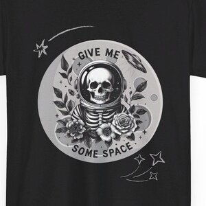 以下が含まれることがあります： 黒いTシャツ。宇宙飛行士のヘルメットをかぶった骸骨のグラフィック、バラ、そして「GIVE ME SOME SPACE」の文字が描かれています。デザインはグレーと白の濃淡です。