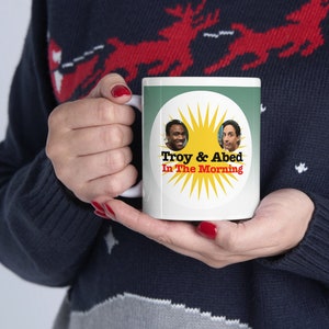 May include: White ceramic mug with a green and yellow design. The design features a sun with two faces inside, and the text "Troy & Abed In The Morning".