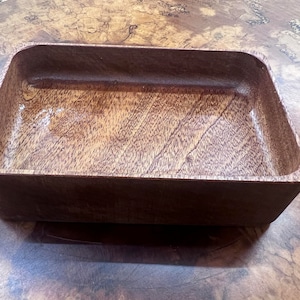 May include: A rectangular wooden bowl with rounded corners. The bowl is made of dark brown wood with a visible grain pattern. The interior has a glossy finish, and the bowl sits on a wooden surface.