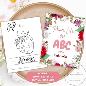 Könnte beinhalten: Zwei weiße Seiten mit Text und Illustrationen liegen auf einem Holztablett. Eine Seite hat eine Erdbeerillustration und das Wort "Fresa". Die andere Seite hat den Text "Primer Libro del ABC para Gabriella", umgeben von einem floralen Rand. Der Text "INBEGRIFFEN Größen - 12,7x17,8cm 20,3x29,2cm BONUS Table Sign" ist ebenfalls sichtbar.