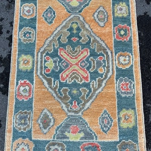 Puede incluir: Una alfombra vintage con un patrón geométrico azul y naranja. La alfombra tiene una forma de diamante central con un diseño floral y está rodeada por un borde de formas geométricas más pequeñas.