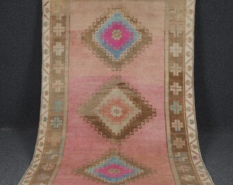 Puede incluir: Una alfombra turca vintage con fondo rosa y tres medallones en forma de diamante en tonos marrones, azules y rosas. La alfombra tiene un borde beige con un patrón geométrico repetitivo.