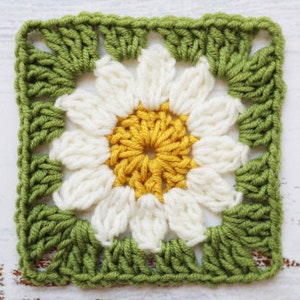 May include: A crocheted square featuring a white daisy flower in the centre. The flower has a yellow centre and green leaves around the edges.